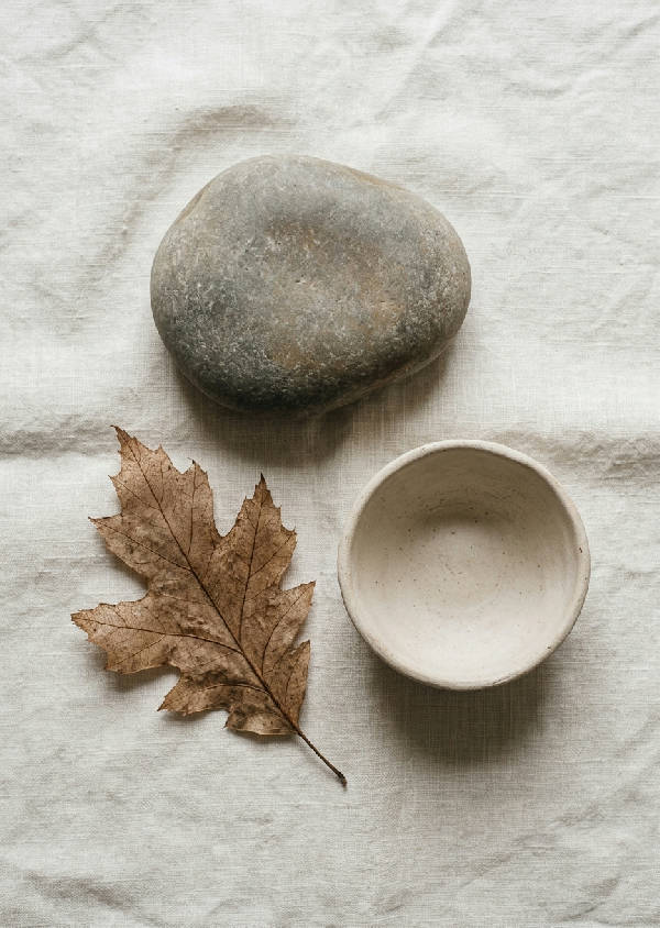 En lugn flat lay med en slät flodsten, ett skört torkat löv och en liten oglaserad keramikskål på ljust tyg.