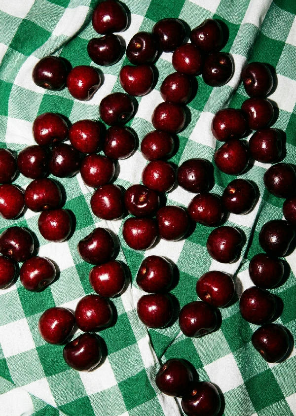 Närbild på glänsande mörka körsbär som vilar på en skrynklig grön och vit rutig picknickfilt, med skarpa skuggor från direkt blixt.