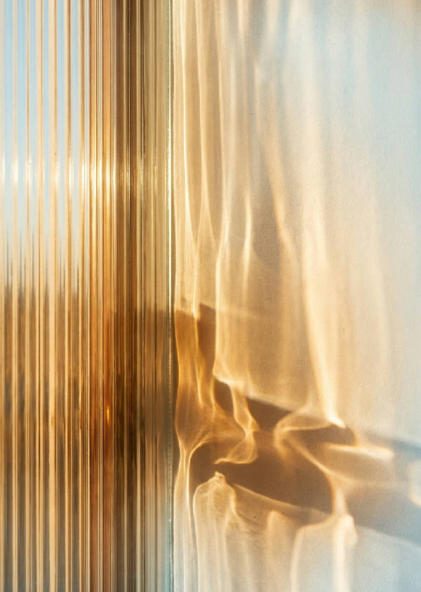 Abstrakt fotografi av solljus som filtreras genom räfflat glas, med förvrängda ljusstrålar och mjuka skuggor på en slät vägg i varma toner.
