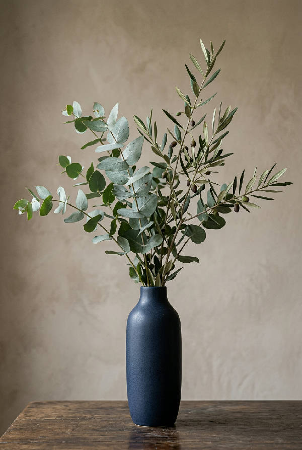 Närbild av eukalyptus- och olivkvistar i en matt svart vas på en varm beige bakgrund med mjuka skuggor. Elegant och minimalistisk still life.