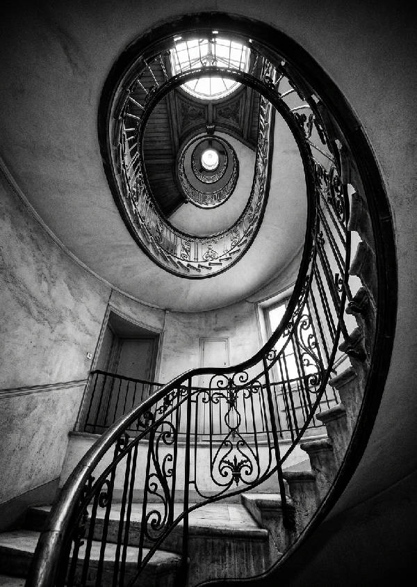 Svartvitt fotografi som visar en spiraltrappa sedd underifrån, detaljerade trappsteg och räcken som skapar ett virvlande mönster uppåt.