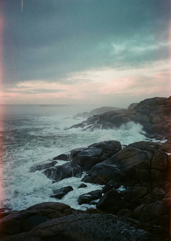 Disig, kornig filmfotografi av nordisk kust i skymning. Vågor mot mörka granitklippor med havsdunkel. Grått, teal, rosa himmel.
