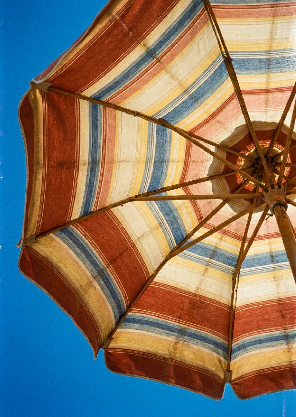 Närbild av ett randigt, solblekt italienskt strandparasoll i azurblått, terrakotta, kräm och gult, fotograferat underifrån mot en molnfri himmel.
