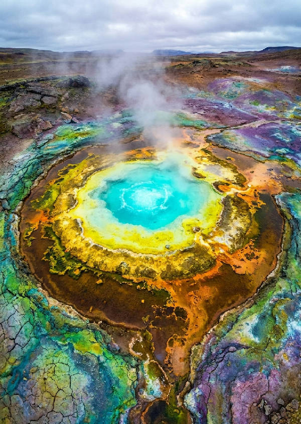 Flygfoto av geotermisk källa. Koncentriska ringar av mineralavlagringar, bubblande turkost vatten och sprucken jord i starka färger.
