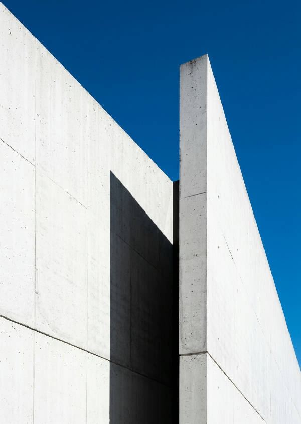 Vertikal bild av en modern, minimalistisk byggnad med raka linjer och skarp skugga mot en klarblå himmel.