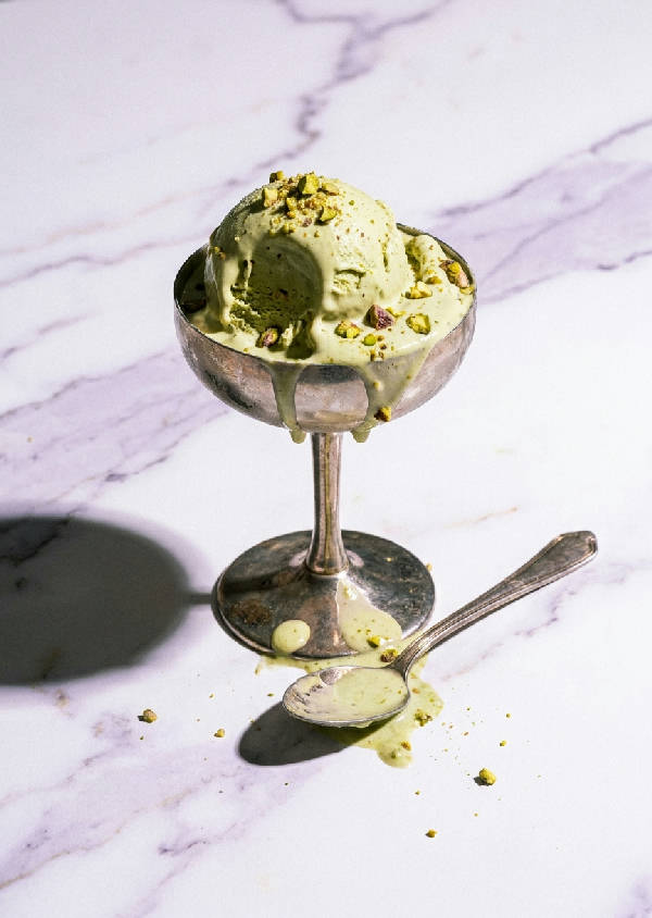 Högmode stillben med halvsmält pistagegelato i silver coupe-glas, silversked på marmor, skarpa skuggor.