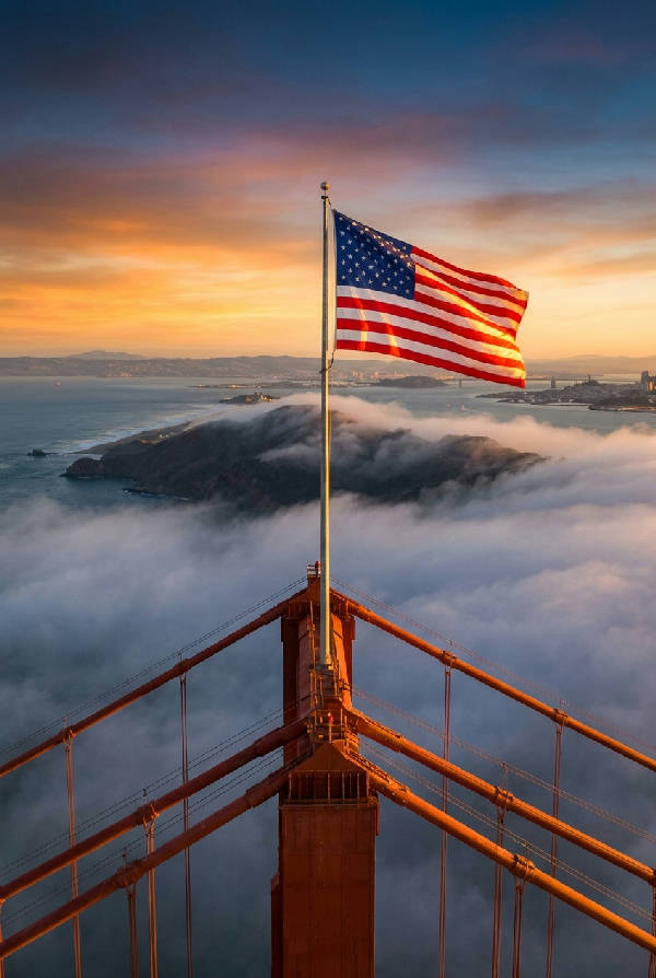 Hyperrealistisk, vertikal bild av Golden Gate-bron vid soluppgången, med amerikanska flaggan vajande högt, dimma som rullar in från Stilla havet. Varmt, gyllene ljus träffar brokablar och flagga.