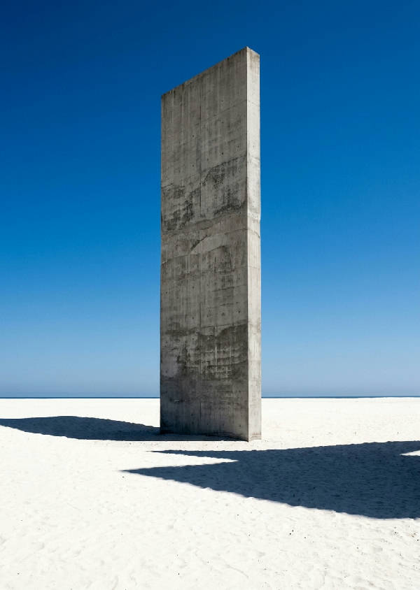 Arkitektoniskt fotografi av en minimalistisk, monolitisk rå betongstruktur på en vit sandstrand under en klarblå himmel.