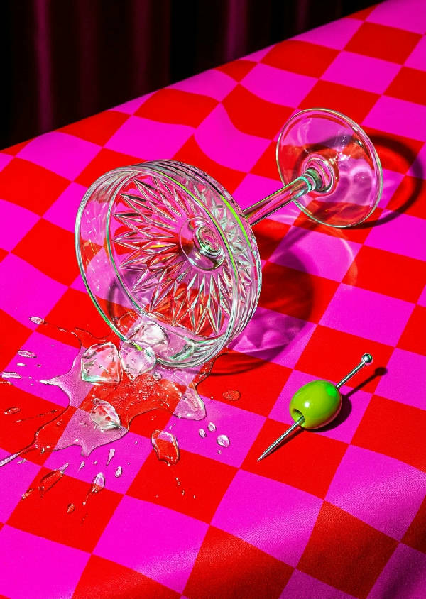 En modern popkonstillustration av en utspilld martini från ett kristallglas med en grön oliv på en tandpetare på en rutig duk. Hög kontrast, dynamisk komposition med dramatisk blixtbelysning.