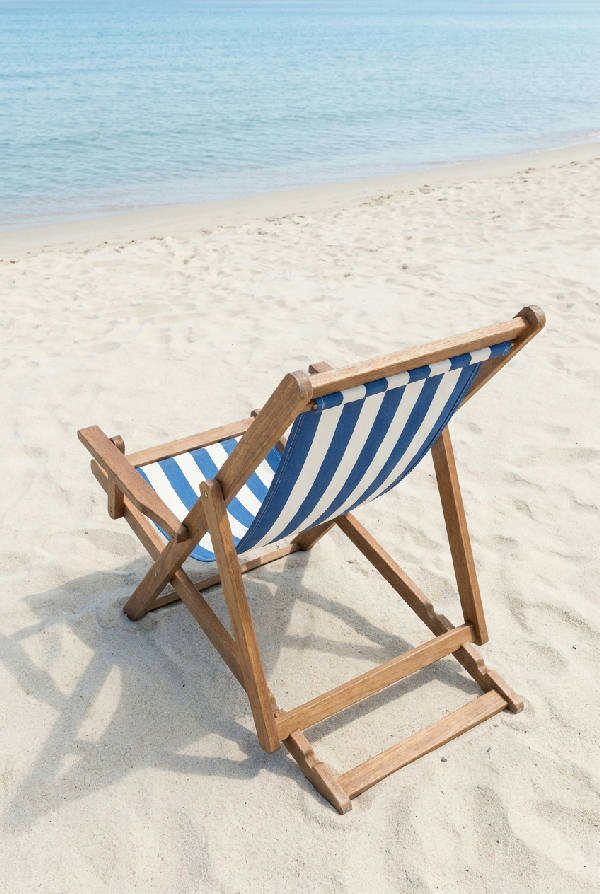 En ensam blå- och vitrandig solstol i trä och tyg, placerad på en ljus sandstrand. Stolen är tom och vänd mot ett lugnt, ljusblått hav med mjuka skuggor i klart dagsljus.