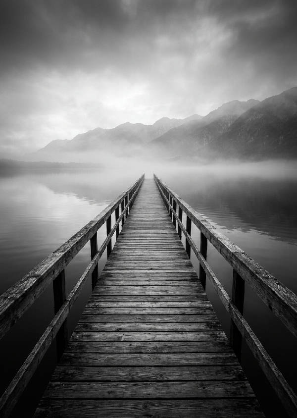 Svartvit minimalistisk landskapsfotografi av en lång träbrygga som leder ut i en lugn dimmig sjö med avlägsna berg i bakgrunden, symmetrisk komposition