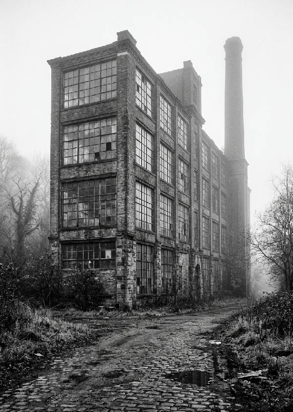 Svartvitt fotografi av en gammal, förfallen industribyggnad i tegel sedd i porträttorientering, med detaljerade texturer och skuggor.