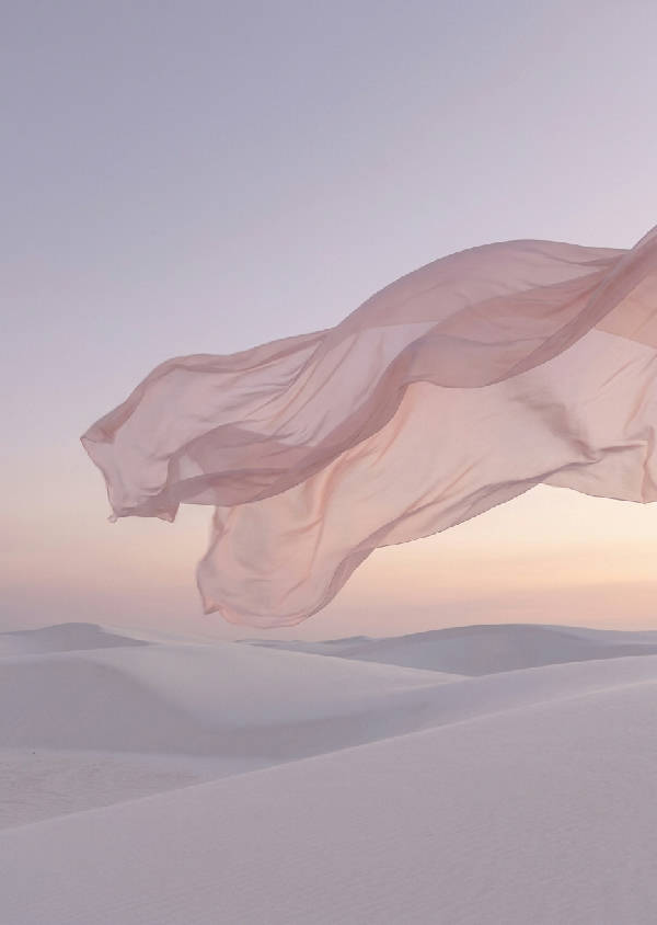 Surrealistisk digitalfotografi av genomskinligt blekrosa siden som svävar över en vit sanddyn under skymningen.