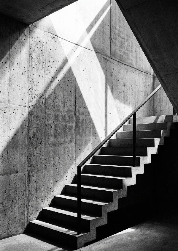 Ett svartvitt fotografi av dramatiska skuggor som faller över en arkitektonisk fasad, skapar abstrakta former och starka kontraster. En minimalistisk komposition i stående format.