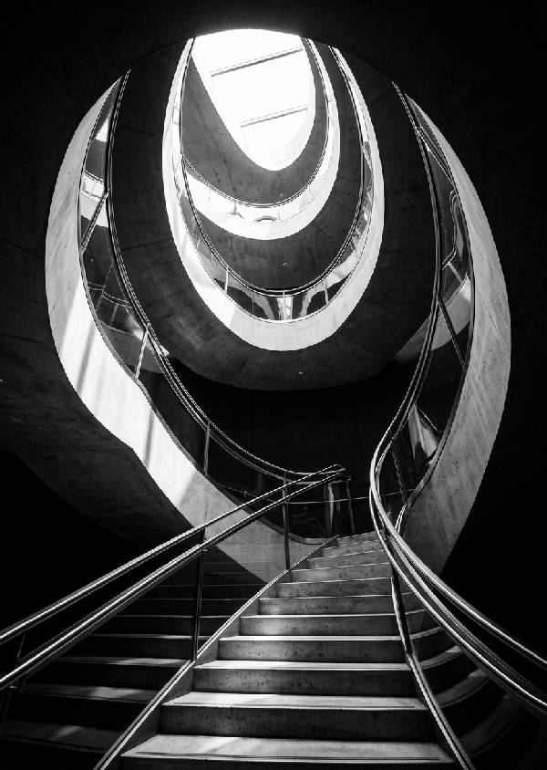 Svartvit fotografi av en dramatisk, modern spiraltrappa sedd underifrån med skarpa linjer och skuggor.
