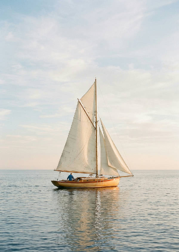 Enkel träsegelbåt med mast och avslappnade off-white segel framför en stillsam marinblå havsyta under en molnfri himmel.