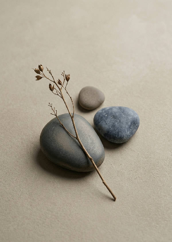 En minimalistisk Japandi-komposition med tre släta, asymmetriska flodstenar och en tunn, torkad gren på en ljus neutral bakgrund.