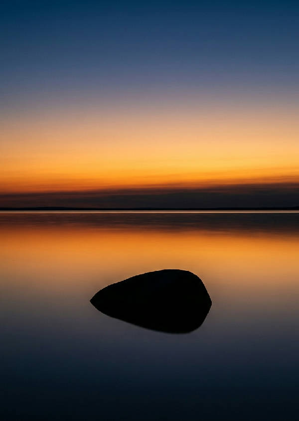 Minimalistisk fotografi av midnattssolen som reflekteras i en spegelblank sjö med en ensam sten i förgrunden.