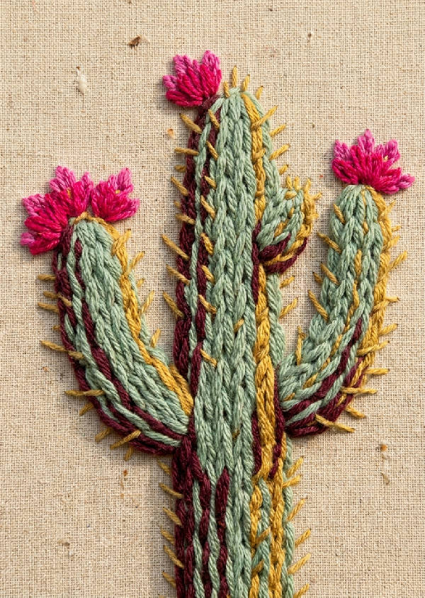 Närbild av en broderad kaktus i ökenblom, med tjocka trådar som skapar en taktil yta av sagegrön, fuchsia och senapsgul på beige bakgrund