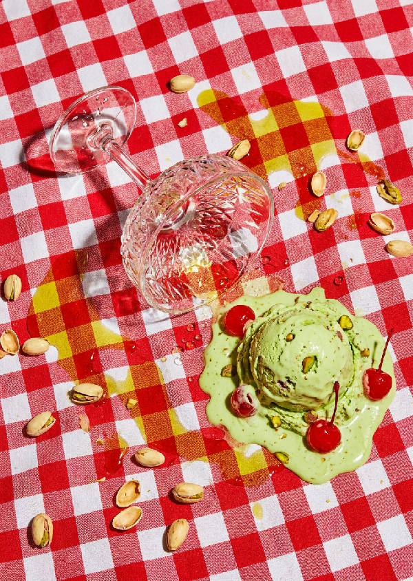 Rörigt sommarbord: smältande pistageglass, röda körsbär, spillt glas, rutig duk. Hög kontrast.