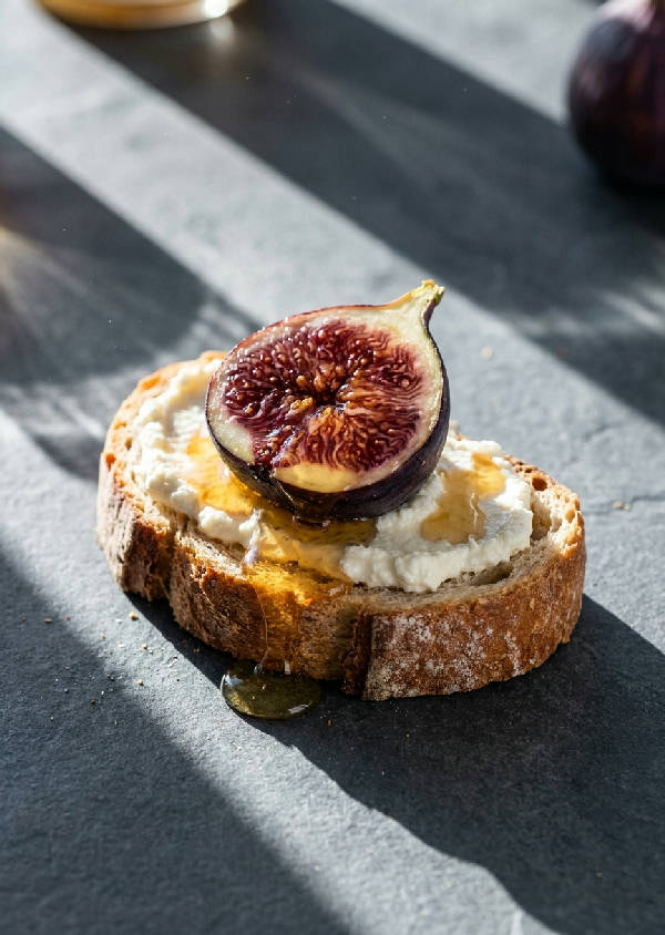 Närbild på en skiva rustik ricotta toast med ett skivat färskt fikon som dränks i gyllene honung, skarpa diagonala skuggor från morgonsolen.