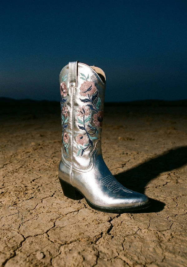 En silvermetallic cowboy boot med blommiga broderier står på sprucken ökenjord vid skymning, fotograferad underifrån med hård blixt.