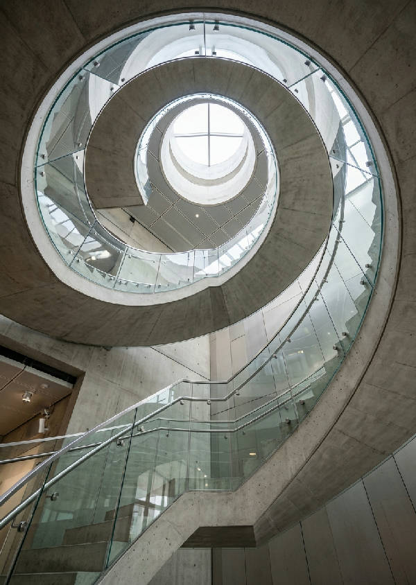 Vertikal bild av en modern, minimalistisk spiraltrappa sedd underifrån, med geometriska former och gråskala.
