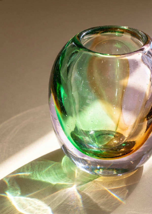 Närbild av en tjock glasvas i porträttorientering som bryter solljus till färggranna ljusrefraktioner i smaragdgrönt, varmt bärnsten och mjuk lila på en beige yta.