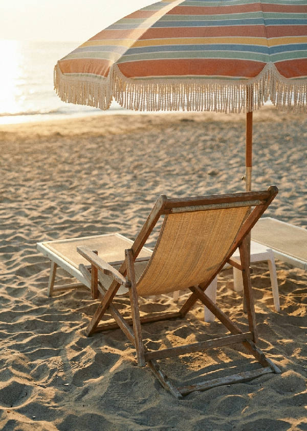 Vintage 1970-tals fotografi av en tom solstol under ett randigt parasoll på en sandstrand i Medelhavet med mjukt eftermiddagsljus och långa skuggor