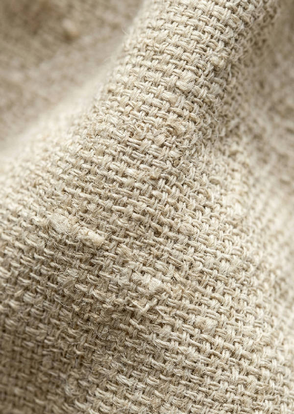 Närbild på en vävd tygtextur i naturfärgad ecru, troligen linne eller bomull, med oregelbunden struktur och mjuk belysning.