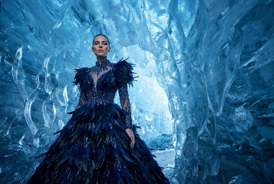 En supermodell i en extravagant midnattsblå fjäderklänning står i en gigantisk glödande iskave med dramatiskt blått ljus.