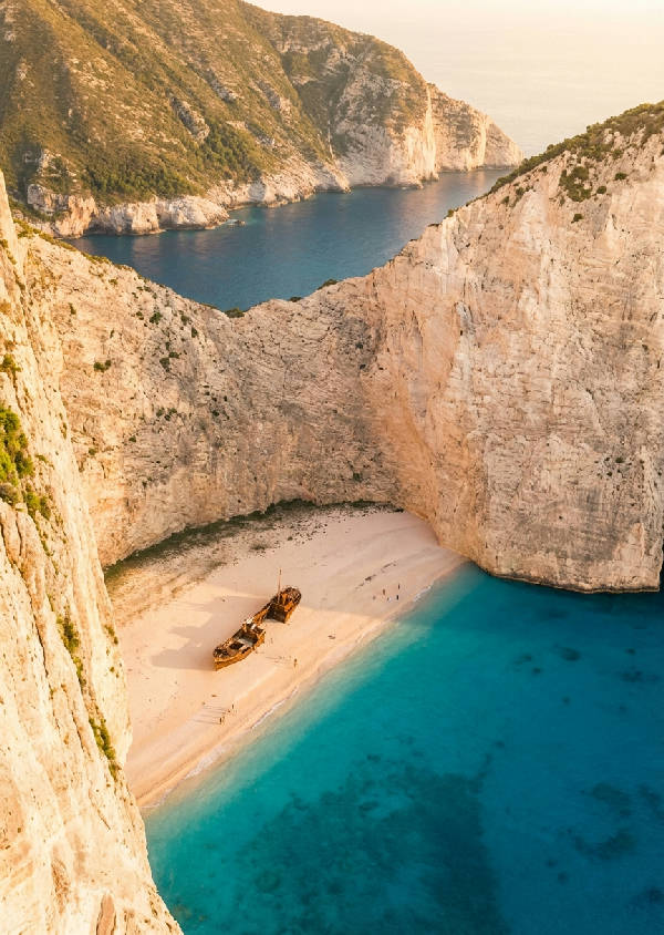 Hyperrealistiskt fotografi av Navagio Beach med ett skeppsvrak inbäddat mellan höga vita klippor och turkost vatten i solnedgången, med långa skuggor och några besökare på stranden.
